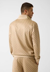 Pullover sportivo beige con motivo a logo ripetuto, collo alto, polsini a costine e vestibilità rilassata, realizzato in tessuto morbido.
