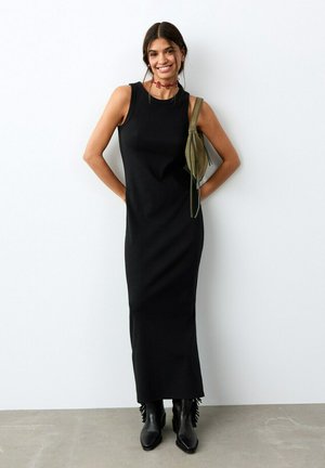 Lächelnde Frau in ärmellosem schwarzem Maxikleid, schwarzen Stiefeln mit Fransen, rotem Halsband und kleiner olivgrüner Umhängetasche steht vor einer weißen Wand.
