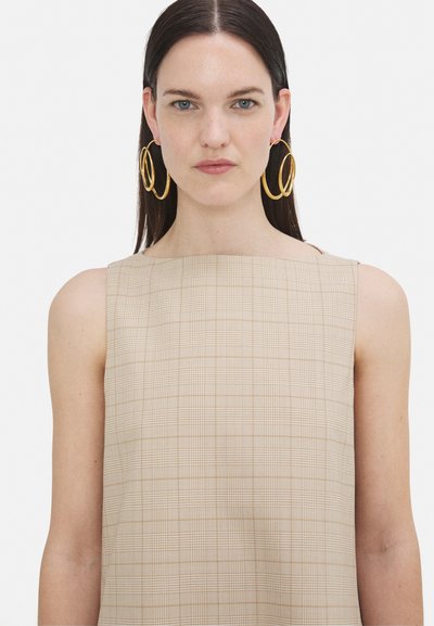Beige karierte ärmellose Bluse mit rechteckigem Ausschnitt; das Modell trägt große goldene Ohrringe. Glattes Material mit einem subtilen Gittermuster.