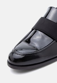 Κοντινό πλάνο ενός γυαλιστερού μαύρου δερμάτινου μοκασίνι με ευρύ μαύρο υφασμάτινο λουρί στο πάνω μέρος και λεπτομερή ραφή κατά μήκος των άκρων.