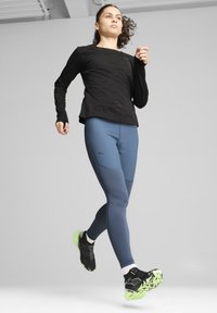Fekete, hosszú ujjú sportfelső, testhezálló kék leggings texturált panelekben, és fekete futócipő neon zöld talppal, dinamikus futó pózban.