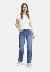 Weißes T-Shirt mit kurzen Ärmeln und Herzdesign, kombiniert mit hellblauen weiten Jeans und silbernen Sneakers, vor einem weißen Hintergrund stehend.