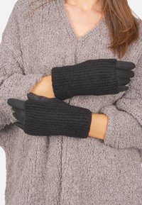 Schwarze gestrickte Handschuhe mit einem gerippten Muster und offenen Fingerspitzen, getragen über einem grauen, flauschigen Pullover. Sichtbare Nähdetails an den Bündchen.