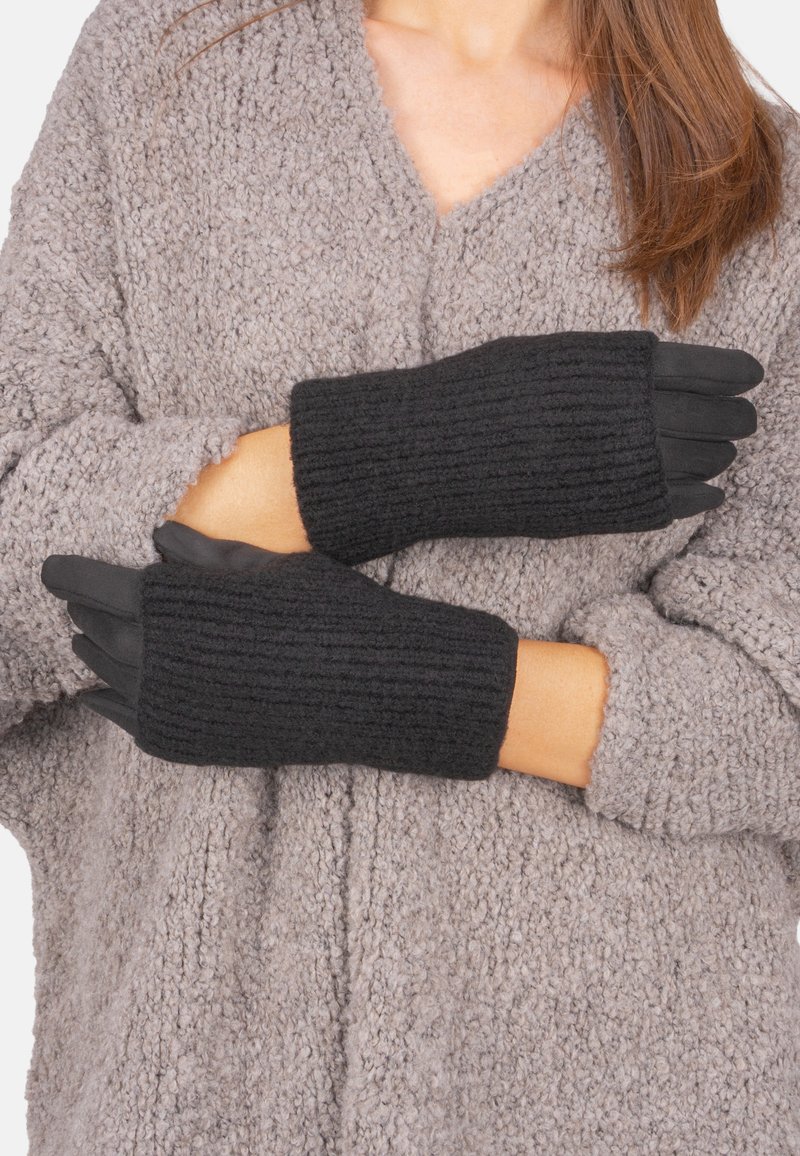 Schwarze gestrickte Handschuhe mit einem gerippten Muster und offenen Fingerspitzen, getragen über einem grauen, flauschigen Pullover. Sichtbare Nähdetails an den Bündchen.