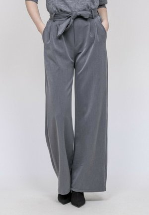 Pantalon droit gris à jambes larges avec ceinture nouée, porté avec un pull gris rentré et des chaussures noires pointues, mains dans les poches.