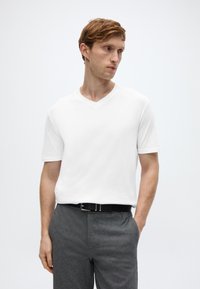 Λευκό T-shirt με κοντά μανίκια και V-λαιμόκοψη, σε συνδυασμό με γκρίζα υφασμάτινα παντελόνια με υφή. Το σύνολο περιλαμβάνει μια μαύρη ζώνη με ασημένια αγκράφα. Μινιμαλιστικός σχεδιασμός.