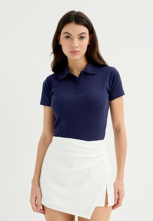 Polo de manga corta azul marino con un acabado texturizado y cuello, combinado con una falda envolvente blanca con una abertura lateral.