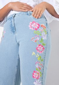 Světle modré džíny z denimu mají pestré květinové výšivky v růžové, červené, žluté a zelené barvě po celé straně. Standardní design se pěti kapsami.