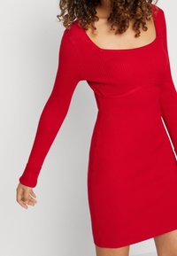 Abito rosso a coste con scollatura quadrata e maniche lunghe, silhouette aderente e lunghezza al ginocchio, realizzato in un materiale a maglia elasticizzato.