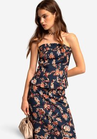 Jonge vrouw in een strapless bloemen-top en bijpassende rok, met een kleine beige satijnen handtas, die naar de zijkant kijkt.