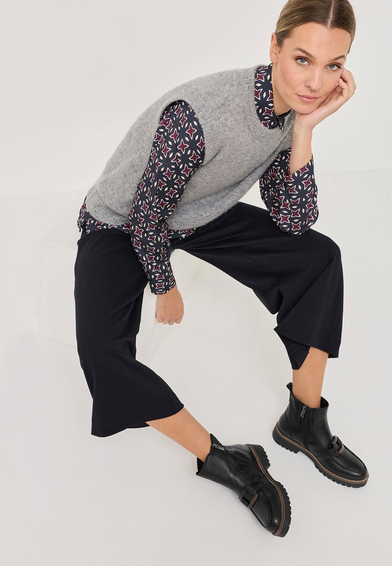 Grauer Woll-Sweater ohne Ärmel über einer blau gemusterten Bluse, kombiniert mit schwarzen, knöchellangen Hosen und schwarzen Ankle Boots mit Schnallen-Detail.