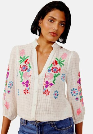 Kvinde iført hvid bluse med farverig blomsterbroderi og 3/4 ærmer, kombineret med blå jeans, mod ensfarvet baggrund.