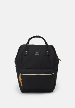 Sac à dos rectangulaire noir avec poignées supérieures, poche avant zippée avec tirette beige, et une petite étiquette de marque centrée près du haut.