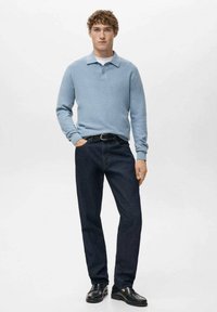 Jasnoniebieska koszulka polo z długim rękawem, ciemne dżinsy i czarne buty, stojące na neutralnym tle. Sweter ma wyraźny kołnierz.