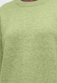 Gros plan d'un pull tricoté vert clair montrant un col rond côtelé et un tissu texturé.
