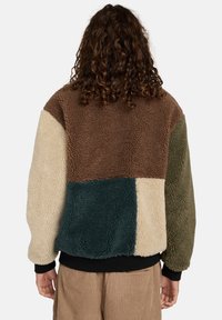 Veste en polaire avec un design patchwork dans des tons de marron, crème, vert et bleu sarcelle foncé, dotée d'un tissu texturé et de poignets côtelés noirs.