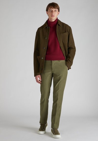 Oliwkowa kurtka button-up noszona na bordowym golfie, w zestawieniu z jasnymi oliwkowymi spodniami i zielonymi trampkami, wszystkie o prostym wyglądzie.