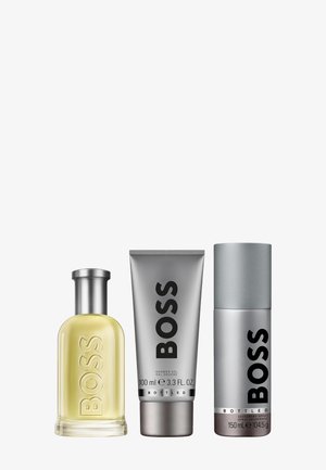 Drie producten: een helder glazen parfumfles met een zilveren dop, een zilveren tube douchegel en een zilveren deodorantspray.