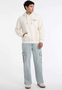 Cremefarbener Hoodie mit einer vorderen Kängurutasche und verstellbaren Kordeln; kombiniert mit hellblauen Cargohosen mit Seitentaschen.