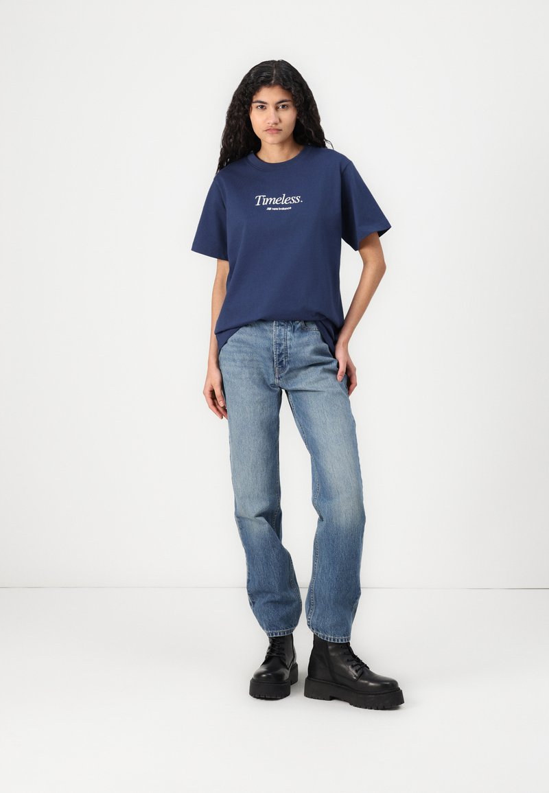 T-shirt en coton bleu marine à manches courtes avec le texte blanc "Timeless.", associé à un jean bleu clair et des bottes plateformes noires.