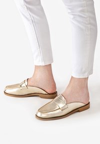 Guldmetalliska slip-on loafers med en slät yta, platt brun sula och en diskret remaccent framtill, tillsammans med vita byxor.