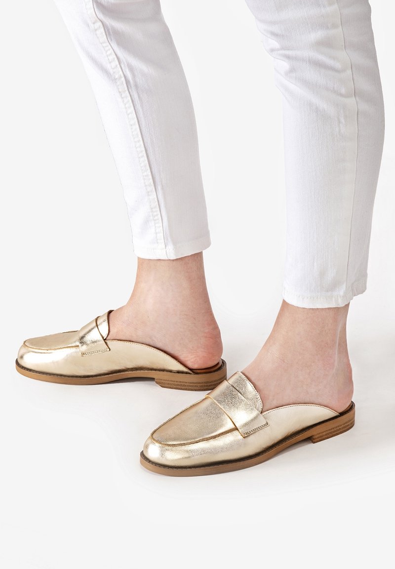 Guldmetalliska slip-on loafers med en slät yta, platt brun sula och en diskret remaccent framtill, tillsammans med vita byxor.