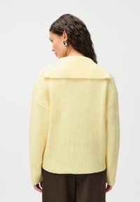 Yellow oversized sweater with a ribbed texture, wide collar, and dropped shoulders. Pairing with brown trousers; back view.