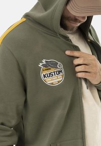 Hoodie vert foncé en mélange de coton, avec des rayures jaunes et un patch rond avec un texte et un design d'aile sur la poitrine.