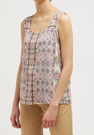 Femme portant un chemisier sans manches à motifs géométriques marron, noir et rouge, associé à un pantalon beige.