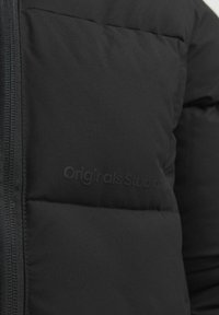 Melnā pufīgā jakā ar gludu tekstūru, ar rāvējslēdzēja aizdari un smalku tonālu logotipu, parādās quilted dizains.