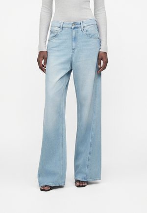 Inimene, kes kannab helesiniseid laia säärega teksapükse, halli pikalehise toppi ja musti avatud varvastega sandaale, seisab valge tausta ees.