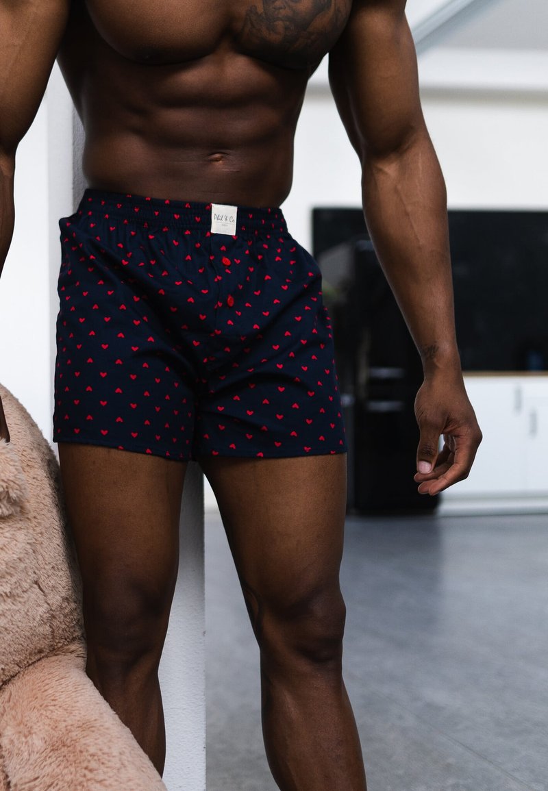 Muskelöser Mann steht drinnen, trägt dunkelblaue Boxershorts mit kleinen roten Herzmustern und einem weißen Etikett am Bund.