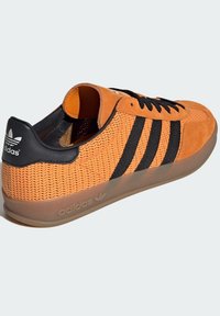 Narančaste suede i mrežaste tenisice s crnim prugama, okruglim vezicama i prozirnom gumenenom potplatom. Ima utisnuti logo na peti.