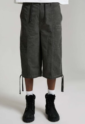 Dunkelgraue knielange Cargo-Shorts mit Kordelzügen am Saum, getragen mit weißen Socken und schwarzen Schnürstiefeln an einer Person vor einfarbigem Hintergrund.
