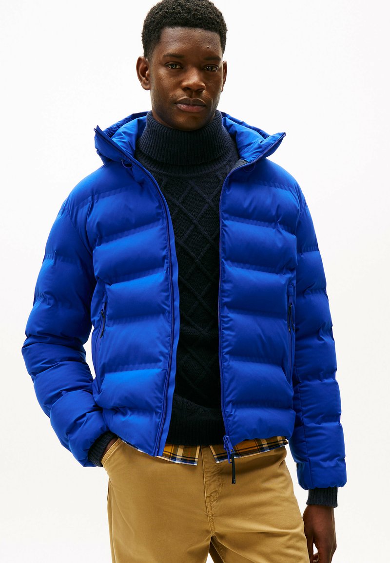Blå puffjakke med et quiltet mønster, der har hætte og lynlåslommer. Båret over en mørk turtleneck og en ternet skjorte.