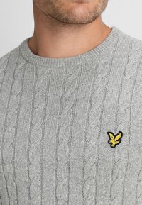 Světle šedý svetr s pleteným vzorem a kulatým výstřihem. Na levé straně hrudi má žluté a černé logo orla. Měkká textura a klasický design.