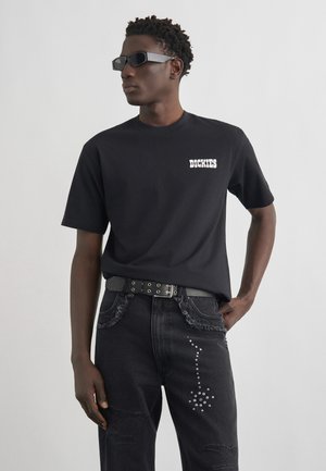 Ung mand iført sort Dickies t-shirt, sort bælte med nitter og sorte jeans med sølvprikket mønster, stående mod en ensfarvet lys baggrund.