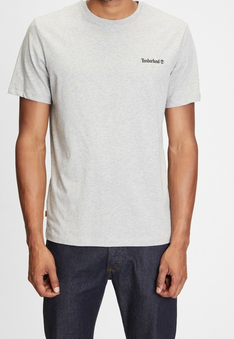 T-shirt en coton gris à manches courtes, col rond, avec un petit logo Timberland imprimé sur la partie gauche de la poitrine. Texture douce.