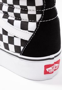 Baskets montantes en toile à motif damier noir et blanc, avec des accents blancs, des panneaux latéraux noirs contrastants et une étiquette de logo rouge au talon.