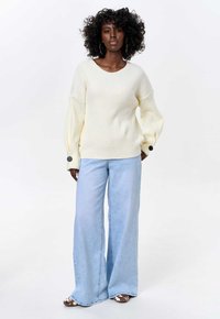 Cream ribbed sweater with wide sleeves and black button details, paired with light blue wide-leg jeans and patterned shoes.