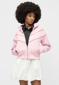 Γυναίκα με μεγάλα σγουρά μαλλιά που φοράει ροζ ζακέτα με φερμουάρ και κόκκινες ρίγες πάνω από μαύρο μπλουζάκι και λευκή φούστα, με τα χέρια στις τσέπες της ζακέτας.