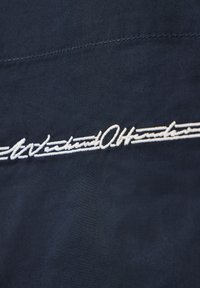 Tissu foncé avec texte cursif brodé en blanc centré horizontalement, orné d'un double soulignement décoratif.