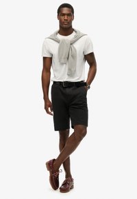 Pantaloni corti neri, t-shirt bianca, maglione grigio annodato attorno al collo, scarpe in pelle marrone con suola in gomma. Posizione in piedi su uno sfondo bianco.