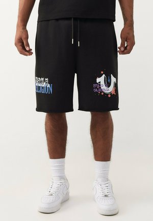 Mann trägt schwarze Shorts mit weißen und blauen Textmustern, weiße Socken und weiße Sneaker, steht vor einem einfachen Hintergrund.
