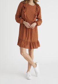 Vestido de color óxido con un escote con volantes y mangas largas, que presenta lunares negros y un dobladillo acampanado. Combinado con zapatillas blancas.