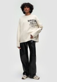 Överdimensionerad krämfärgad sweatshirt med svart grafisk text, kombinerad med svarta byxor i konstläder med vida ben och bruna sneakers.