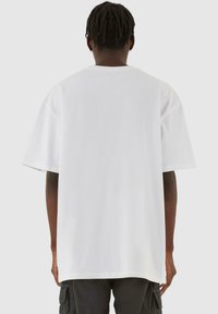 Hvid oversized t-shirt med korte ærmer, rund halsudskæring og en glat tekstur, vist fra ryggen mod en neutral baggrund.