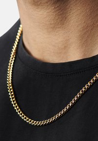 Guld kæde med sammenflettede led, der ligger på en sort skjorte. Kæden ser skinnende ud med en glat overflade.