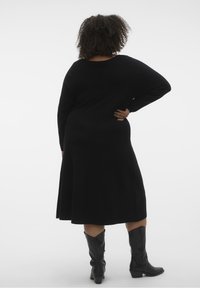 Robe noire en tricot à manches longues et coupe décontractée, avec un ourlet évasé. Portée avec des bottes noires montantes. Texture lisse, design minimal.