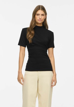 Femme aux longs cheveux bruns portant un haut noir cintré à manches courtes et un pantalon beige ample, debout devant un fond blanc uni.